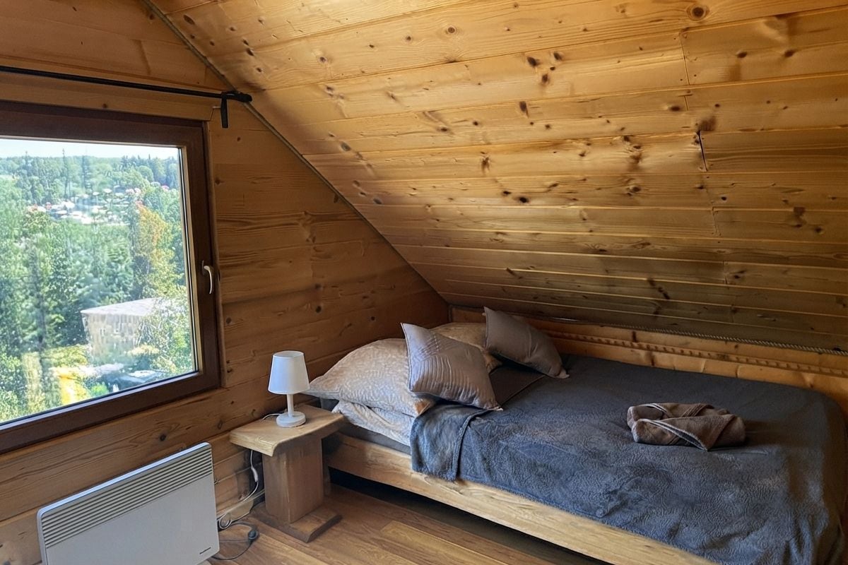 Un dormitori de cabana de fusta amb sostre inclinat, un llit amb almoadines, una tauleta de nit amb una làmpada i vista a l'exterior.