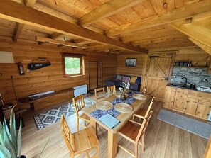 A wooden cabin interior with a dining table, chairs, a sofa, a kitchen area, and a window.