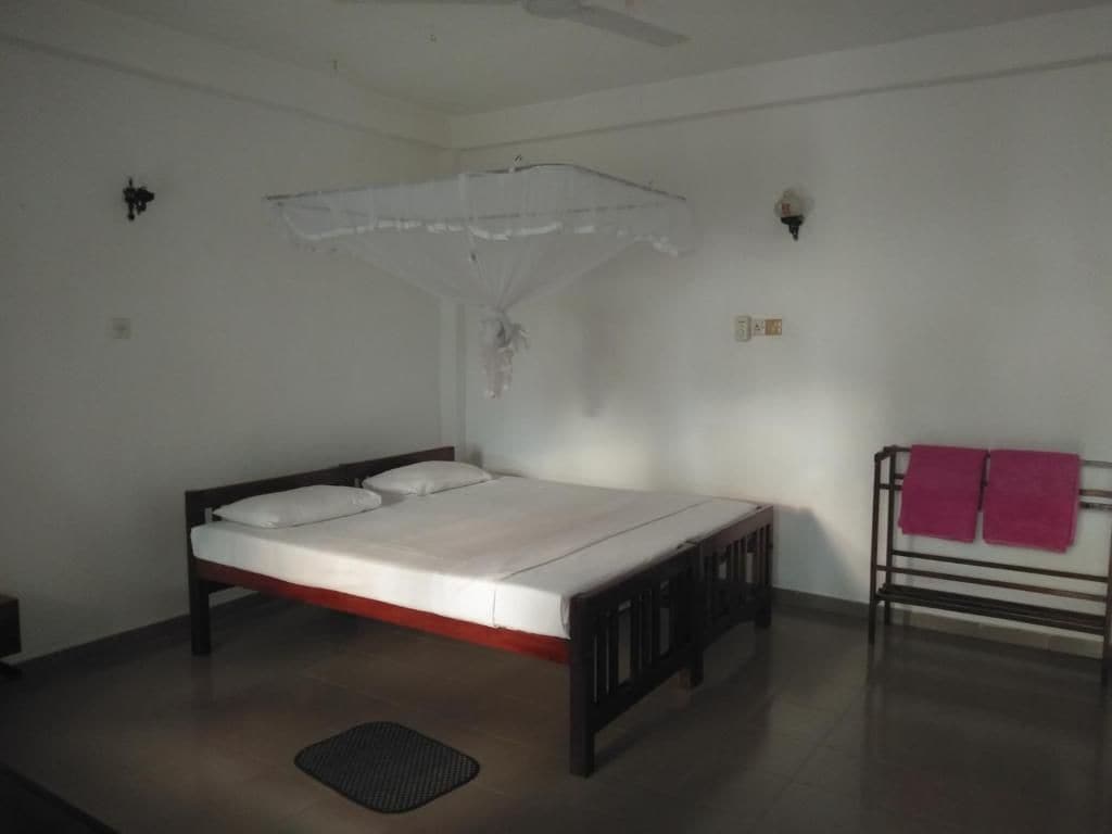 A single bed with a wooden frame and white bedding, a small bedside table, a wall-mounted light fixture, and a ceiling fan.