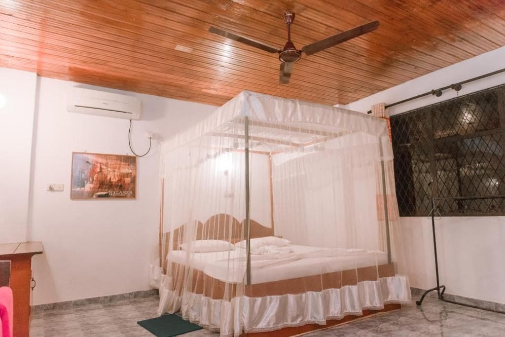 Una habitación con una cama bajo un dosel, un ventilador de techo, un equipo de aire acondicionado, una ventana con reja de metal y un cartel en la pared.