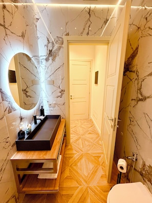 Un baño con pared de mármol, tocador de madera, espejo redondo y un inodoro.