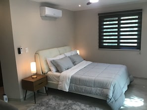 A bedroom with a bed, bedside lamps, a nightstand, an air conditioner, a window with blinds, and a small rug. - LOL-HA House, by the sea—a secluded and peaceful spot (Isla Mujeres)
