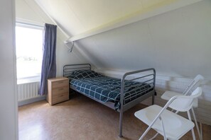 Quarto pequeno, com uma cama de solteiro, mesa de cabeceira de madeira, uma cadeira branca e janela com cortinas.