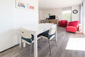 Uma sala de estar moderna com uma mesa de jantar branca, duas cadeiras brancas, um sofá vermelho e uma televisão sobre um aparador de madeira.