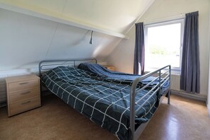A single bed with a metal frame, a bedside table with drawers, a window with curtains, and a sloped ceiling.
