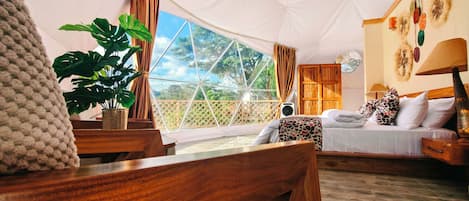 A tented accommodation room with a bed, bedside table, lamp, and a large plant.