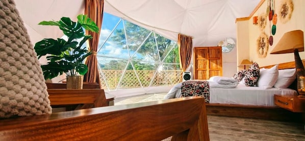Une pièce aménagée comme une tente, avec un lit, un bureau en bois, une plante et une vue sur l’extérieur.