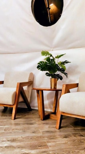 A room with two wooden chairs, a small wooden table with a potted plant, and a round wall-mounted light fixture. - Nekoli Glamping (Arusha)