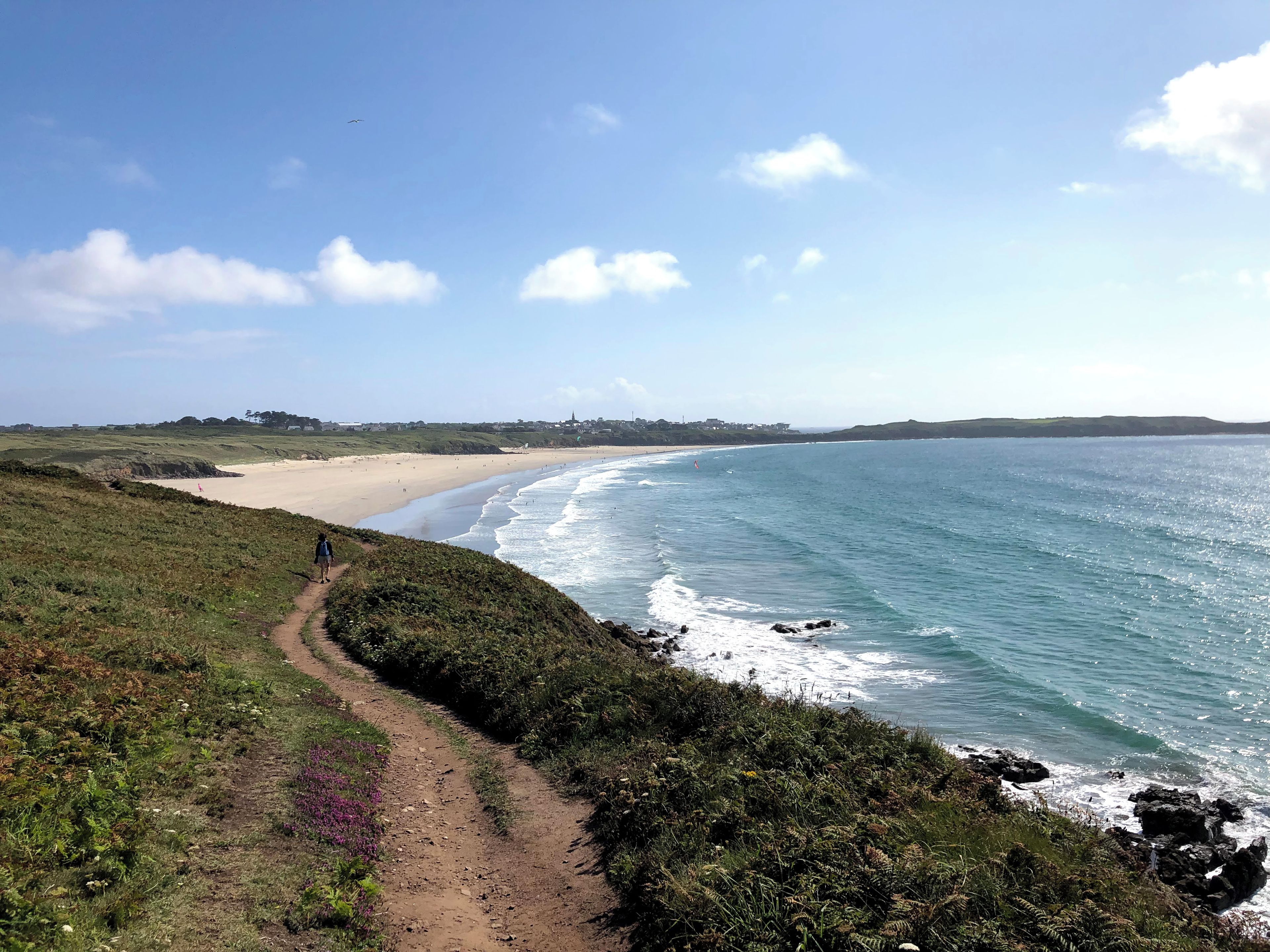 Un sentier côtier qui mène à une plage d’où l’on a une vue dégagée sur l’océan et les collines lointaines.