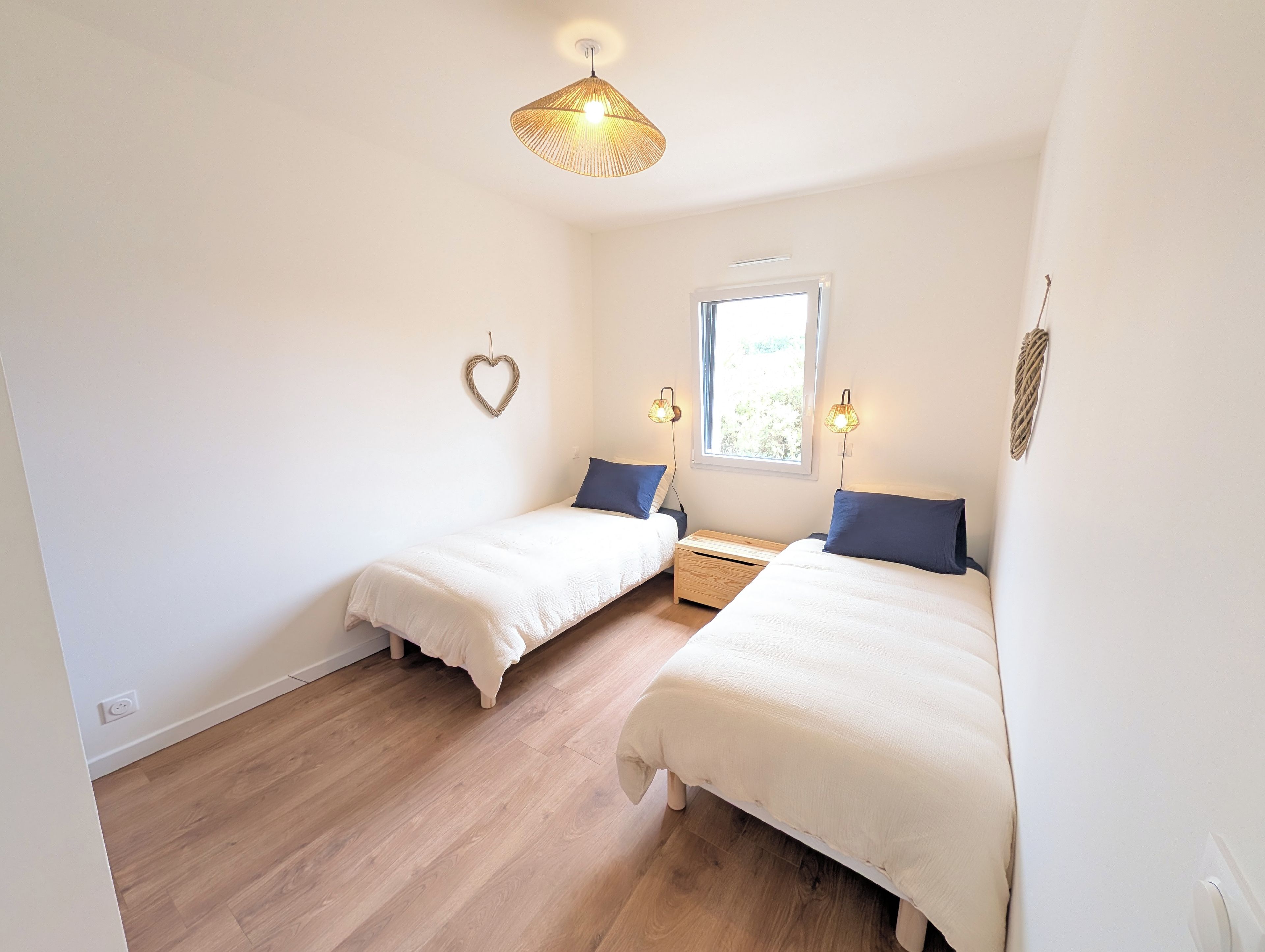 Un dormitorio con dos camas, una ventana, una lámpara colgante y una decoración con forma de corazón en la pared.