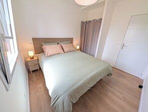 A bedroom with a bed, bedside tables, a nightstand, a window, and a door. - Vrbo Property (Le Conquet)