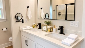 Un baño moderno con un tocador blanco, un espejo grande y una planta en un recinto de vidrio.