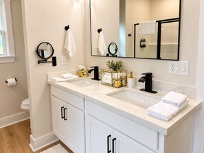 Un baño moderno con un tocador blanco, un espejo grande y una planta en un recinto de vidrio.