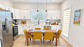 Una cocina moderna con armarios blancos, un refrigerador de acero inoxidable, una mesa de comedor de madera con cuatro sillas y un ventanal con persianas.