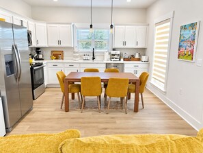 Una cocina moderna con armarios blancos, un refrigerador de acero inoxidable, una mesa de comedor de madera con cuatro sillas y un ventanal con persianas.