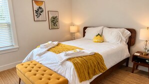 Una cama bien tendida con sábanas blancas y un plaid amarillo, un reposapiés amarillo y dos lámparas fijadas a la pared.