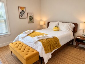 Una cama bien tendida con sábanas blancas y un plaid amarillo, un reposapiés amarillo y dos lámparas fijadas a la pared.