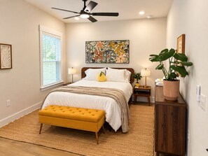 A bedroom with a bed, bedside lamps, a ceiling fan, a window with blinds, a large abstract painting, a potted plant, and a wooden bench. - SunKissed on the Coast, well appointed, high-end home near DT. SMEG Espresso (Pensacola)