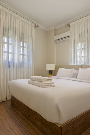 A hotel room with a large bed, white linens, a wooden headboard, a wall-mounted air conditioner, and sheer curtains. - VESTA  - Luxury APT - 2BR - South Teseen (III) (New Cairo City)