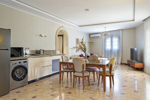 A modern kitchen with a washing machine, a dining area with a table and chairs, a living room with a sofa and a TV, and a large window with curtains. - Luxury APT - 3BR - South Teseen (I) (New Cairo City)