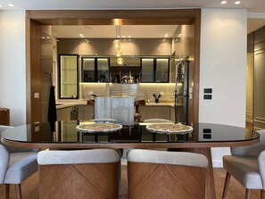 A modern dining area with a glass table and chairs, a central light fixture, and a kitchen in the background. - VESTA  - Luxury APT - 3BR - New Cairo (XXX) (New Cairo City)