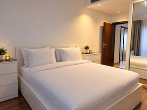A modern bedroom with a large bed, white bedding, a bedside table with a plant, and a mirror on the wall. - VESTA - Luxury Apt- 3br - cfc (xlvii) (New Cairo City)