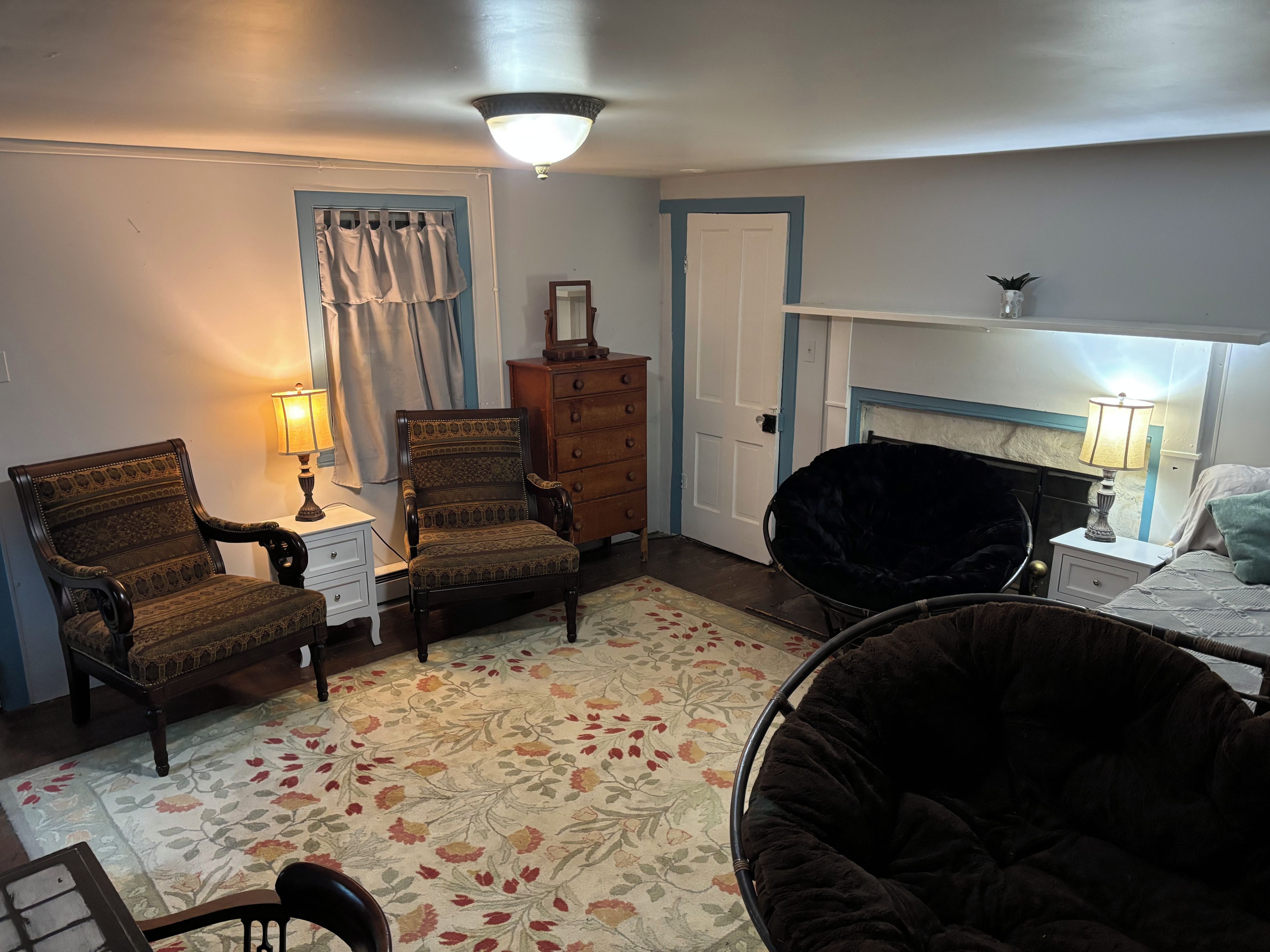 A room with a floral-patterned rug, two armchairs, a black bean bag chair, a dresser, and a bed with a headboard.