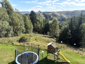 Ein Spielplatz mit Trampolin, Rutsche und einem hölzernen Spielhäuschen, umgeben von einer Rasenfläche und Bäumen.