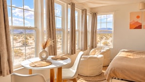 Una habitación con una cama, una mesa redonda con sillas y ventanas grandes con cortinas.