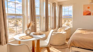 A room with a bed, a round table with chairs, and large windows with curtains.