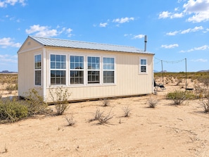 A small, single-story building with a metal roof and multiple windows, situated in a desert landscape with sparse vegetation. - Dark Sky off grid Retreat near Big Bend (Terlingua)