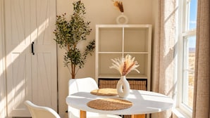 Una habitación con una mesa redonda, dos sillas, una planta en maceta, un florero con flores secas y una ventana con cortinas.
