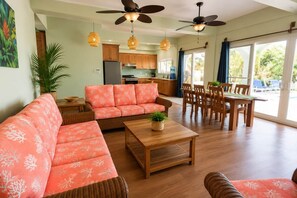 A spacious living room with a sofa, a dining table, and chairs, a kitchen in the background, and large windows with curtains. - Villa Terrapin Station in Hopkins, Beachfront, Private Pool + Rooftop Tiki Bar (Hopkins)