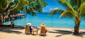 A beach scene with a wooden pier extending into the water, two people seated at a table, and a palm tree to the right. - Villa Terrapin Station in Hopkins, Beachfront, Private Pool + Rooftop Tiki Bar (Hopkins)