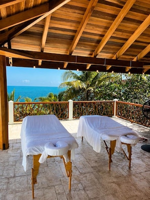 Zwei Massagetische unter einer Holzw pergola mit Blick auf das Meer.