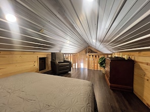 Ein Zimmer mit schrägem Dach, Kamin, Bett, Stuhl und Kommode.
