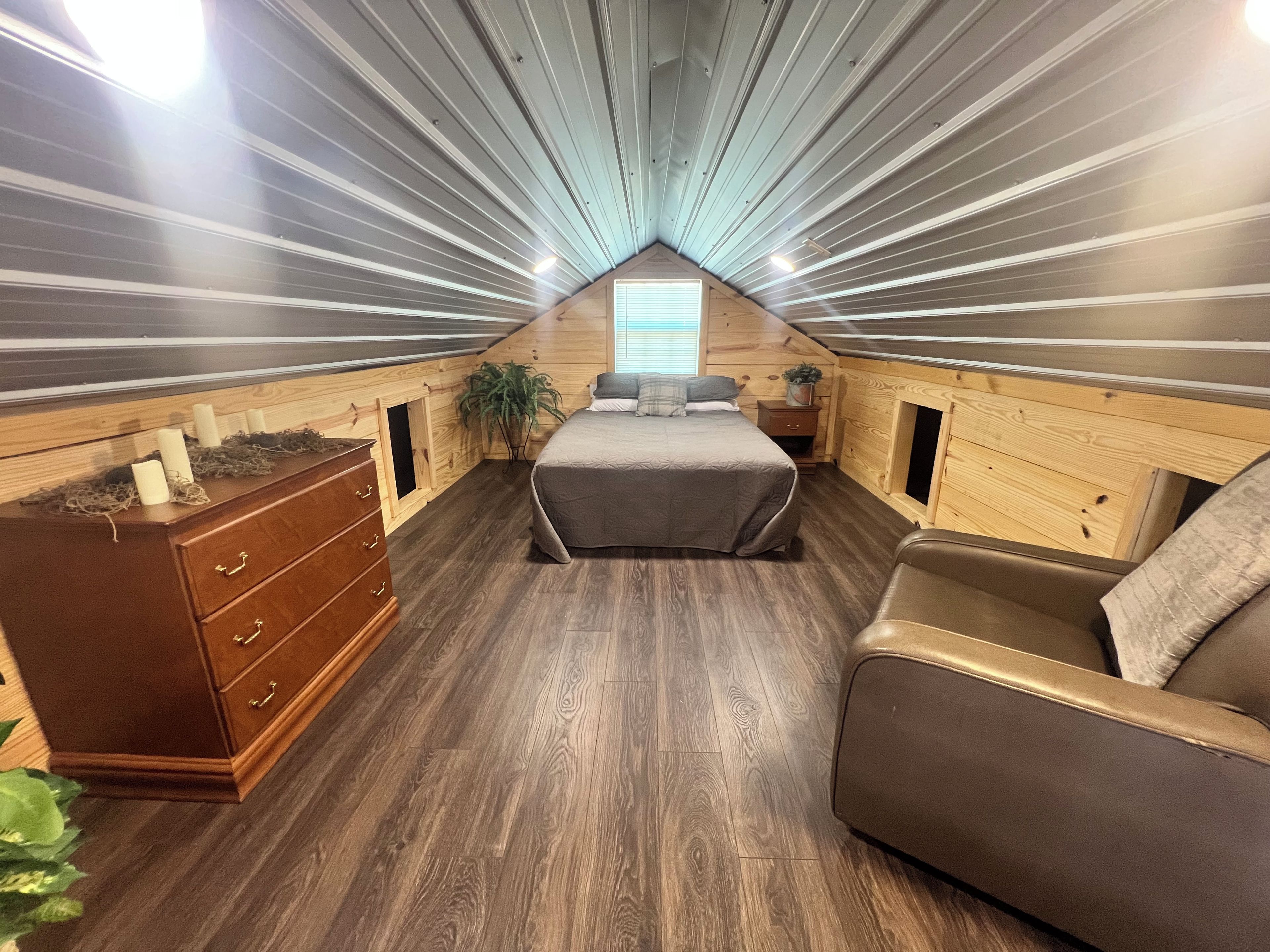 A cozy attic bedroom with a sloped ceiling, wooden walls, a bed, a dresser, and a chair.