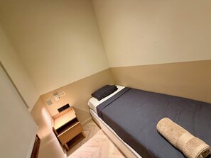 Ein kleines Zimmer mit einem Einzelbett, einem Nachttisch aus Holz, einem Handtuch und einer Wandleuchte.