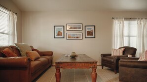 Una sala de estar con un sofá de cuero marrón, una mesa de centro de madera y un sillón marrón. Hay tres cuadros en la pared.