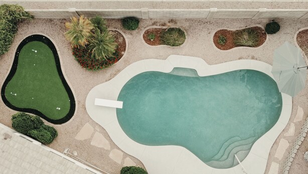 Una piscina con un deck perimetral, un pequeño jardín con diversas plantas y un camino pavimentado que lleva a la piscina.