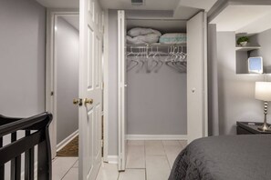 A bedroom with a bed, a bedside table with a lamp, a closet with hanging clothes, and a small shelf with a plant. - Cozy 2 BR Basement Suite (Washington)