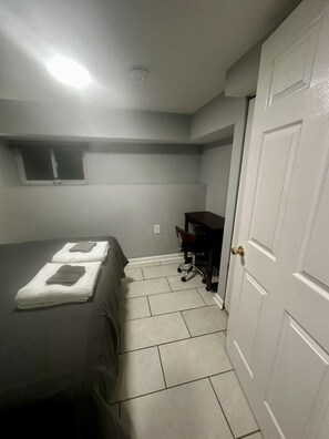 A small, dimly lit room with a single bed, a desk, and a closed door.