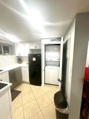 A compact kitchen with white cabinets, a black refrigerator, a washing machine, and a small trash bin. - Cozy 2 BR Basement Suite (Washington)