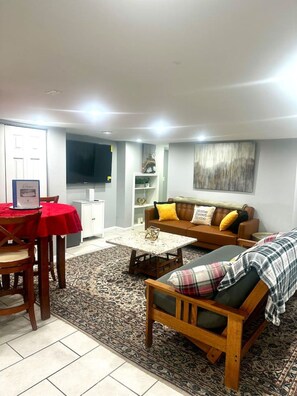 A living room with a sofa, a coffee table, a dining table with chairs, a television, and a bookshelf. - Cozy 2 BR Basement Suite (Washington)