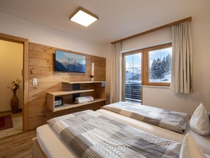 A hotel room with two beds, a mounted TV, a window with a view of snow-covered mountains, and wooden paneling on the walls. - Heart of the Ahornblick House (Brandberg)