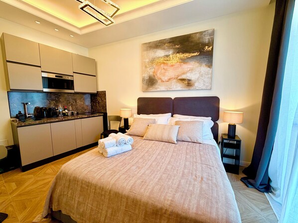 Habitación de hotel con cama, mesitas de noche, un cuadro, zona de cocina y vistas al exterior.