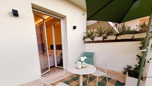 Ein kleiner Patio-Bereich mit Tisch und zwei Stühlen, eine Schiebetür zu einem Schlafzimmer und ein grüner Sonnenschirm, der Schatten spendet.