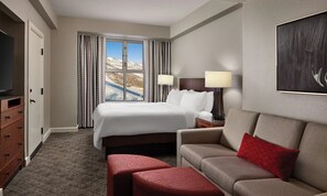 Quarto de hotel com cama, televisão, sofá, um banquinho vermelho e janela com cortinas.