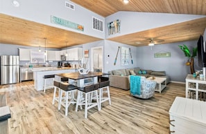 A spacious living area with a wooden ceiling, a kitchen with modern appliances, and a dining area with a wooden table and white chairs. - Beach House 3Bed/2Bath with Ocean View & Golf Cart (Surfside Beach)
