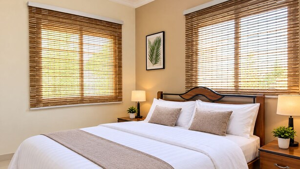 Quarto de hotel com cama, duas mesas de cabeceira, um criado-mudo, planta emoldurada e janelas amplas com persianas.